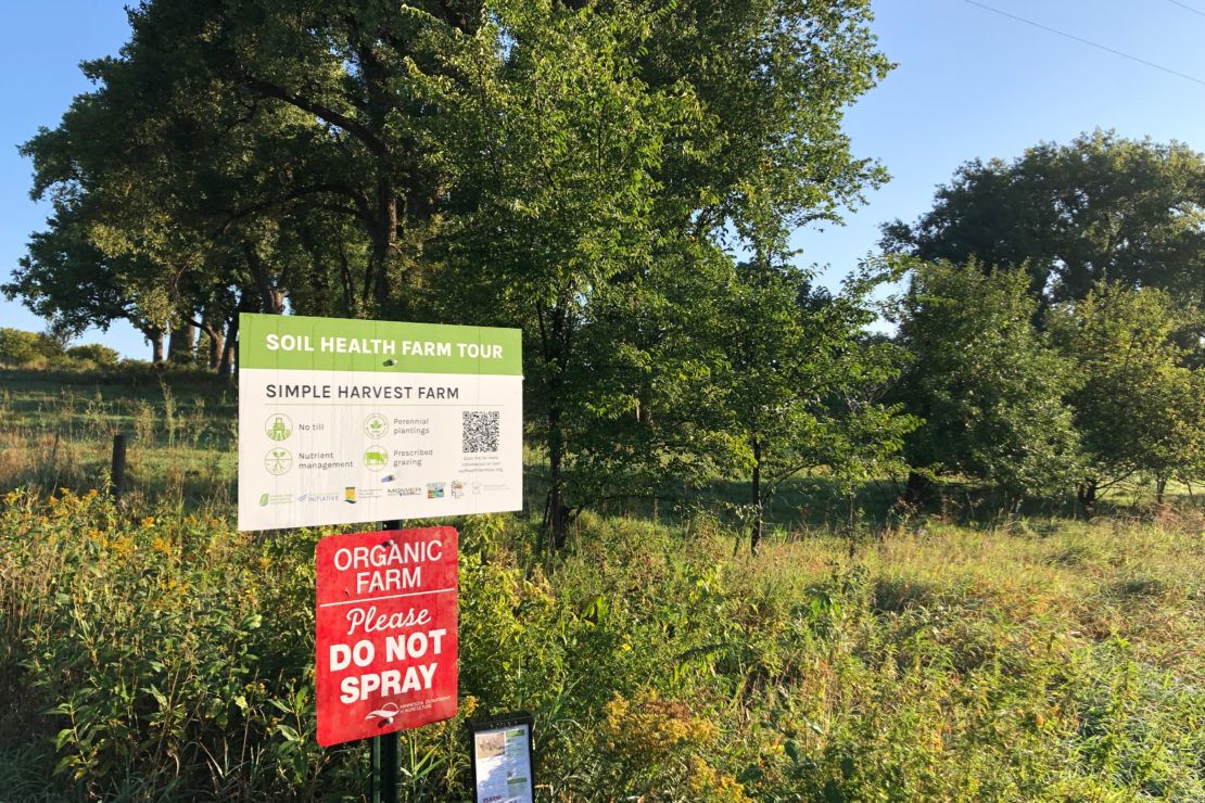 Sign reading soil health farm tour on a post in front of apple trees on a summer day.