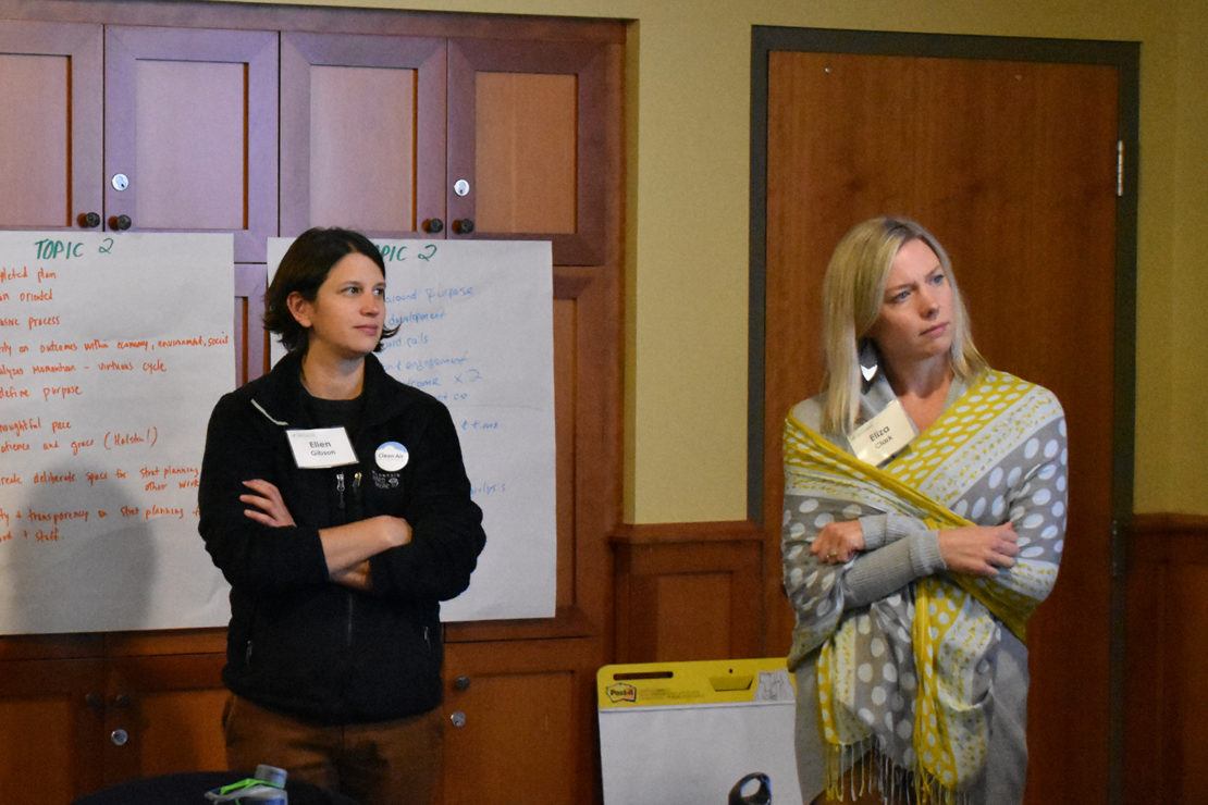 two caucasian women listen to a conversation in a room with easels and notepads in the background