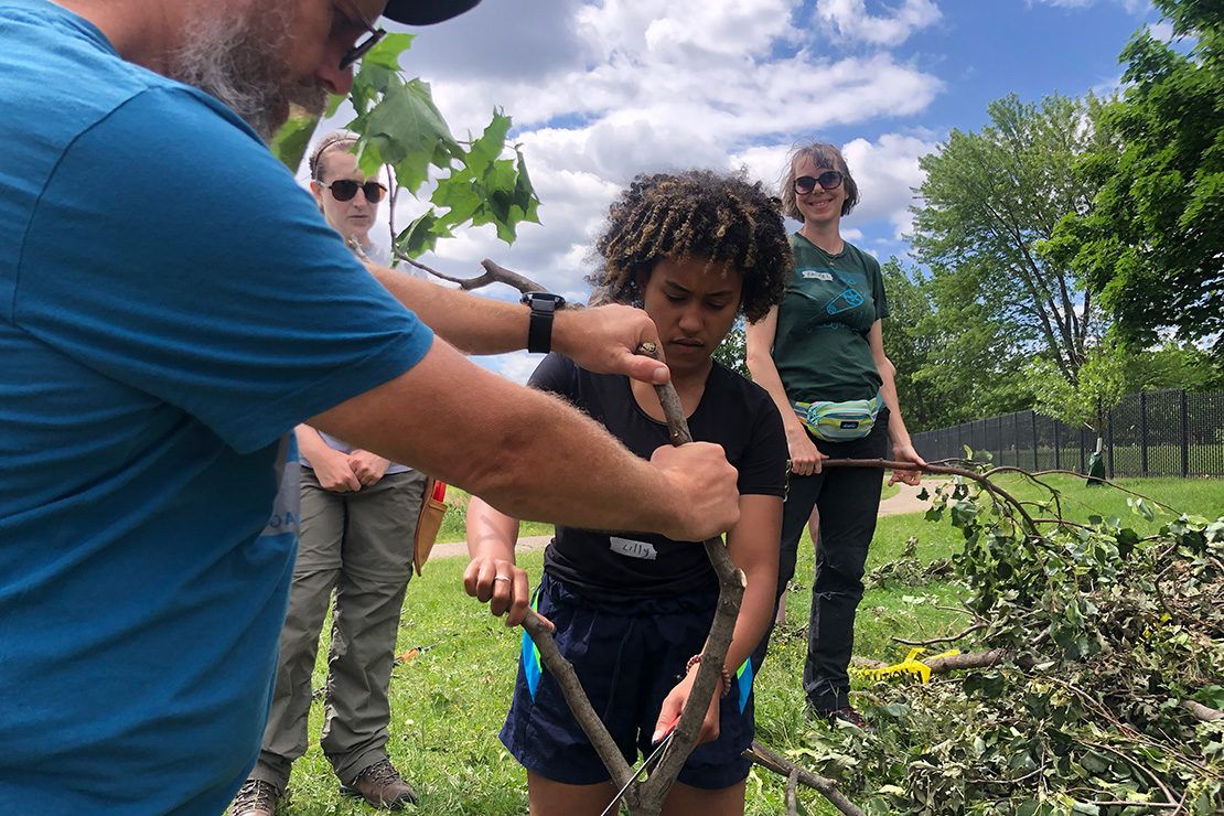Lily Hood holding small saw and trimming tree branch with assistance from man wearing blue shirt.