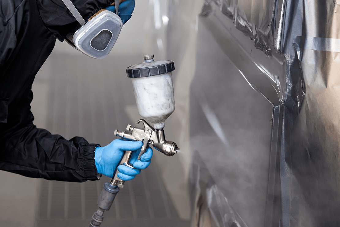Person wearing black coveralls, respirator, and gloves holding a paint gun and painting a van with black paint