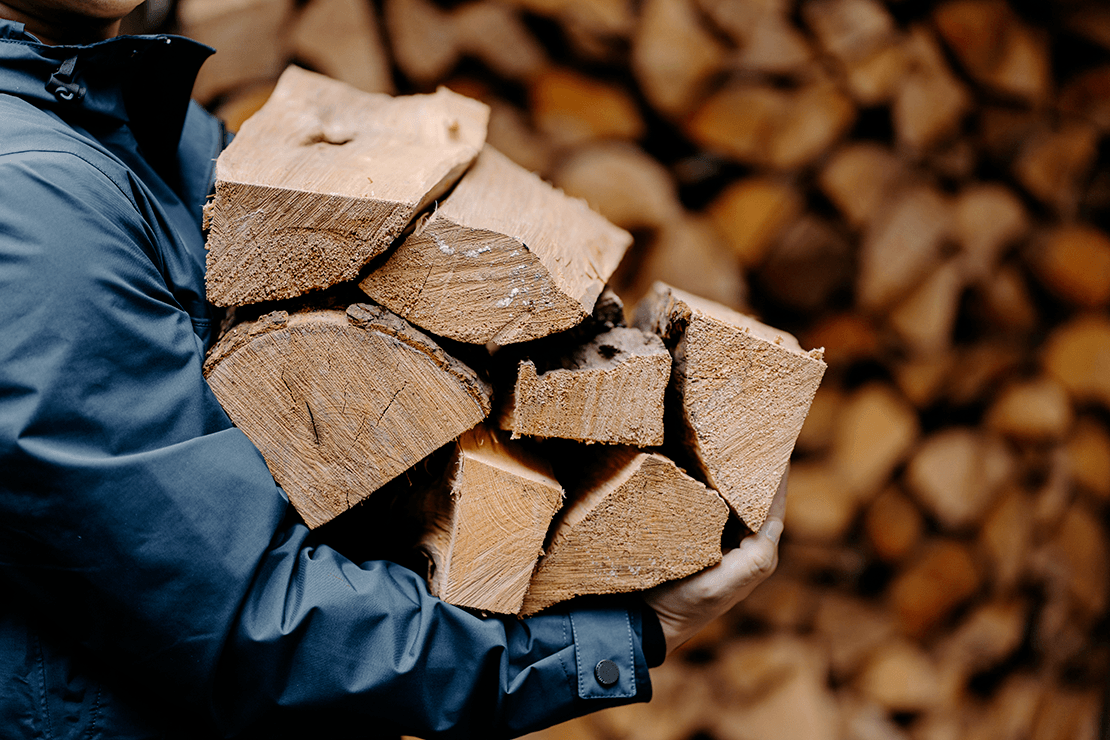 Person wearing a dark blue jacket carrying seven pieces of wood. A pile of wood is visible in the background.