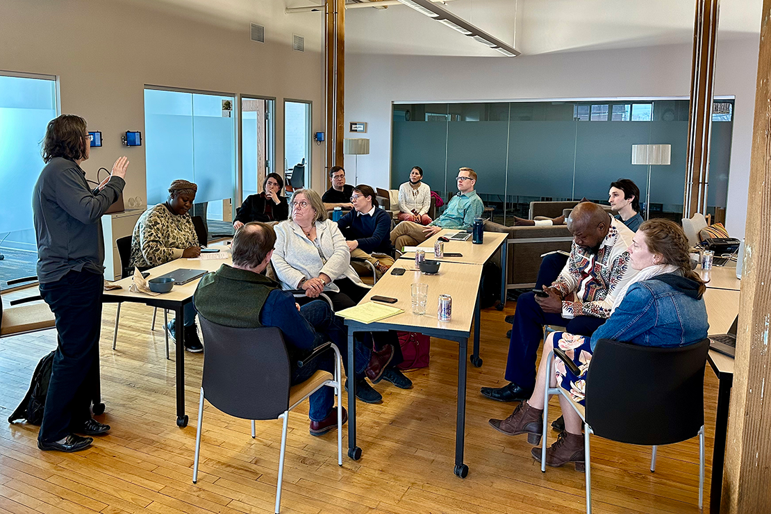 A presenter stands at the left gesturing while speaking to a diverse group of attentive adults seated at tables arranged in a horseshoe formation in a bright, modern meeting room with large windows and a high ceiling. The participants are focused on the speaker, with some taking notes and others listening intently, fostering an atmosphere of engagement and collaboration.