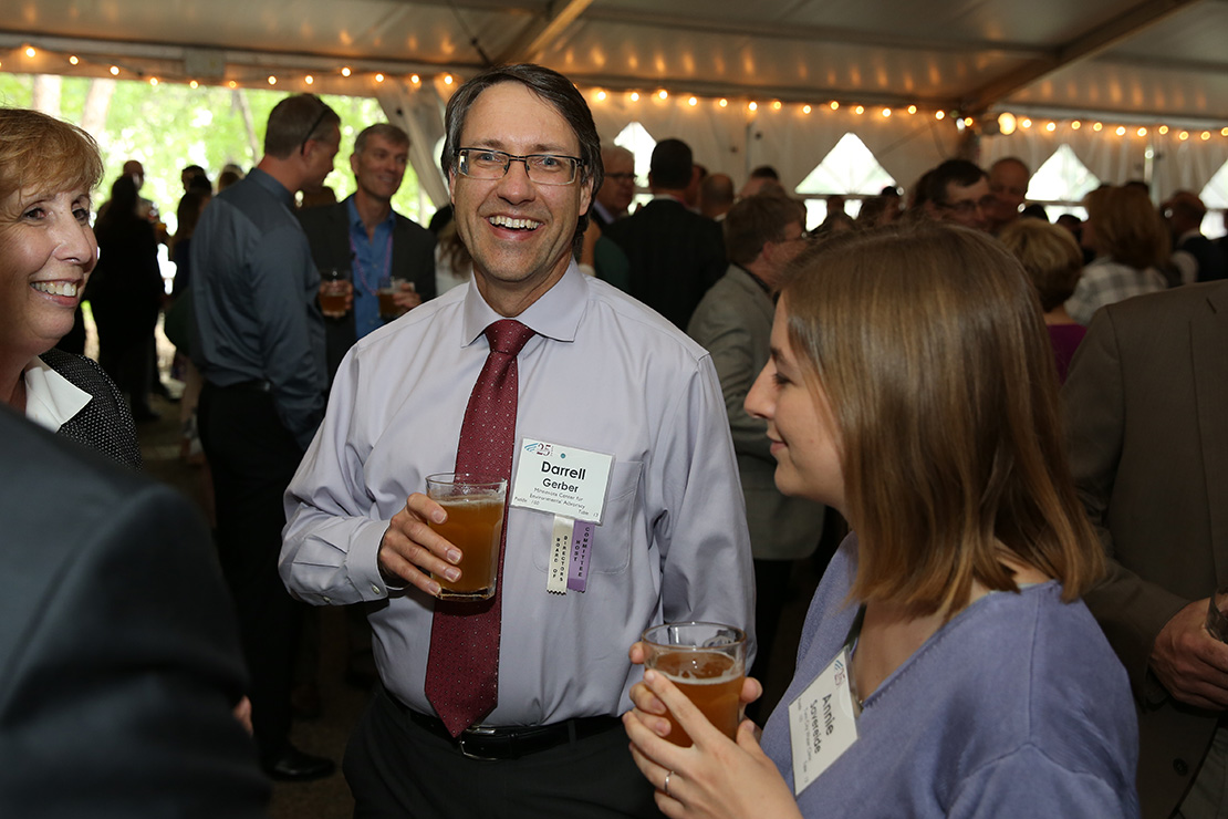 Darrell Geber at the Environmental Initiative Awards reception. He is wearing glasses, a light purple shirt, and a red tie. He is smiling while holding a glass of beer. A woman in a light purple sweater is standing next to him, also holding a glass of beer.